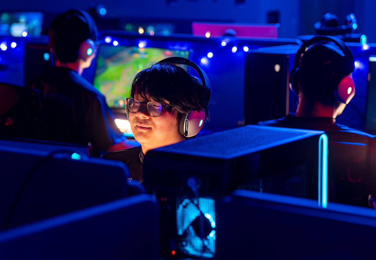 A person wearing headphones and glasses is focused on a screen in a dimly lit room with blue lighting, surrounded by other individuals also engaged in gaming.